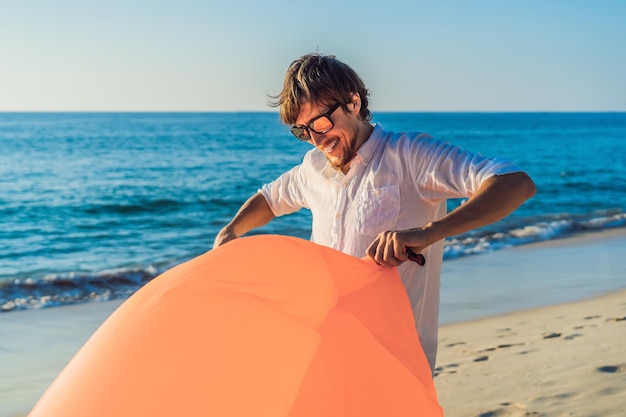 Portrait de style de vie d'été de l'homme gonfle un canapé orange gonflable sur la plage de l'île tropicale Se détendre et profiter de la vie sur un lit d'air