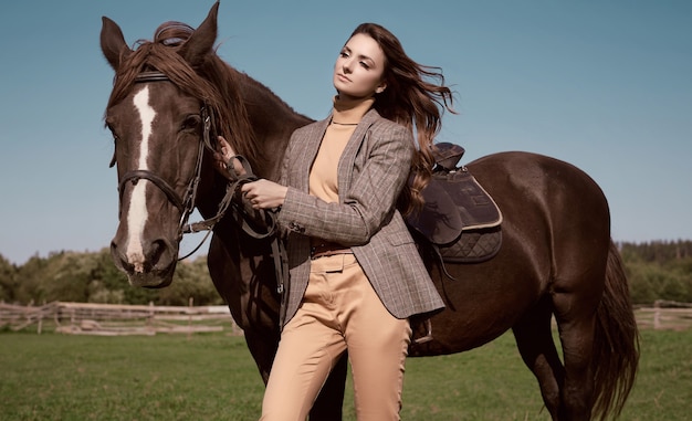 Portrait d'une superbe femme brune dans une élégante veste marron à carreaux posant avec un cheval sur paysage de campagne