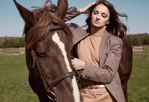 Portrait d'une superbe femme brune dans une élégante veste marron à carreaux posant avec un cheval sur paysage de campagne