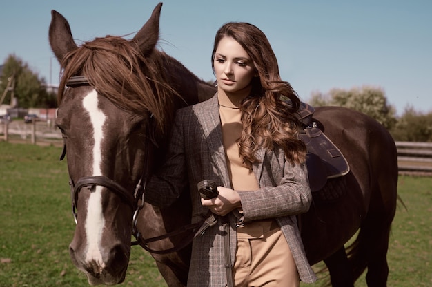 Portrait d'une superbe femme brune dans une élégante veste marron à carreaux posant avec un cheval sur paysage de campagne