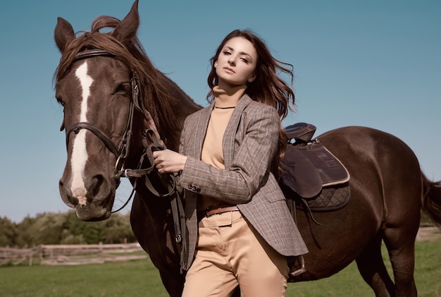 Photo portrait d'une superbe femme brune dans une élégante veste marron à carreaux posant avec un cheval sur paysage de campagne