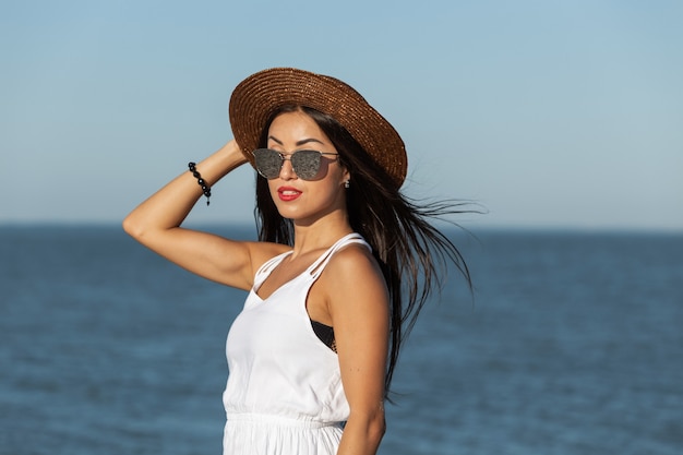 Portrait d'une superbe fille brune en robe blanche, lunettes de soleil et chapeau marron près de la mer par une journée ensoleillée. .