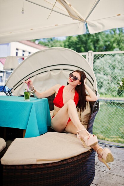 Portrait d'une superbe fille en maillot de bain bikini rouge et lunettes de soleil profitant de son temps en s'asseyant sur le canapé sur le quai au bord du lac