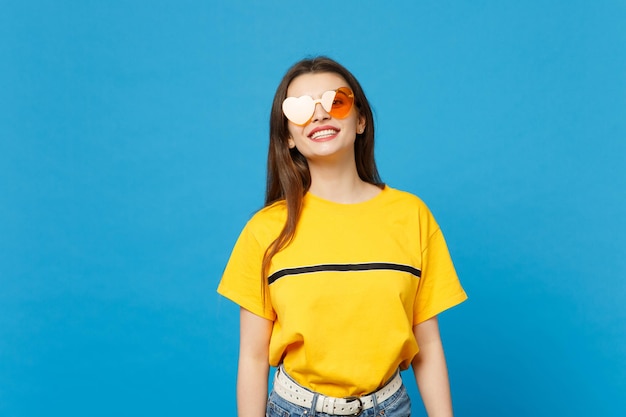 Portrait d'une superbe jeune femme souriante dans des vêtements décontractés vifs, lunettes de coeur debout, regardant la caméra isolée sur fond de mur bleu en studio. Concept de style de vie des gens. Maquette de l'espace de copie.