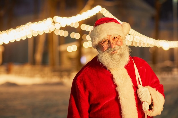 Portrait à la taille du sac de transport du père noël souriant avec des cadeaux la nuit avec des guirlandes lumineuses à ba