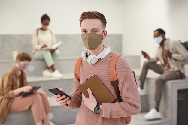 Portrait à la taille d'un étudiant moderne portant un masque et tenant un sac à dos tout en posant dans le salon de l'école en regardant la caméra, espace pour copie