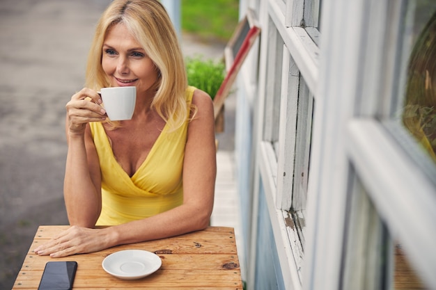 Portrait de taille d'une femme d'âge moyen rêveuse souriante avec une tasse de boisson assise à l'extérieur