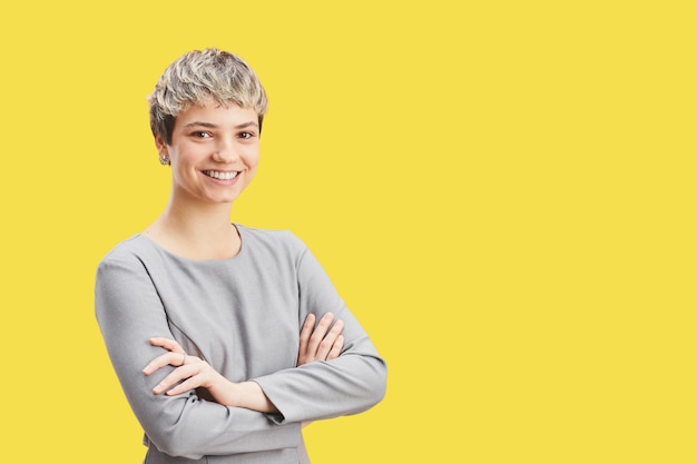 Portrait de taille d'une femme confiante aux cheveux courts souriant à la caméra tout en se tenant les bras croisés sur fond jaune pop, espace pour copie