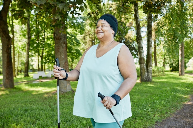 Portrait à la taille d'une femme noire mature souriante marchant avec des bâtons dans le parc pendant l'entraînement en plein air