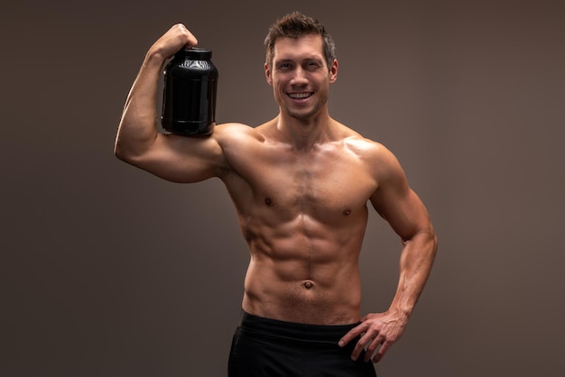 Portrait à la taille d'un homme torse nu sportif posant devant le bocal de l'appareil photo avec une boisson saine, des protéines de lactosérum isolées sur fond de studio marron