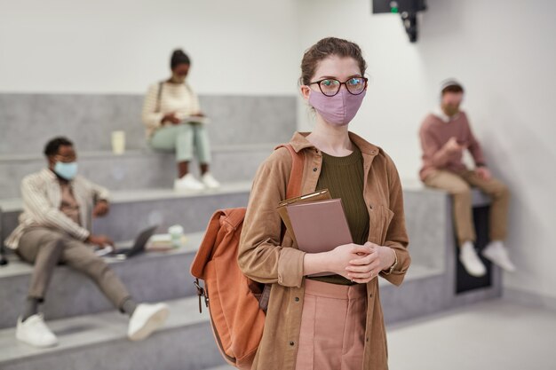 Portrait de taille d'une jeune étudiante portant un masque et regardant la caméra en se tenant debout dans le salon de l'école moderne, espace pour copie