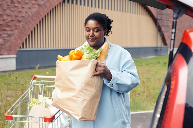 Portrait à la taille d'une jeune femme noire mettant un sac d'épicerie dans le coffre d'une voiture sur le parking d'un supermarché