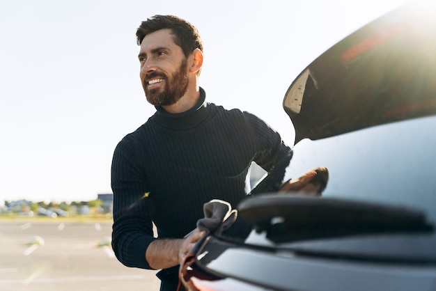 Portrait de taille d'un jeune homme de race blanche nettoyant sa voiture avec une microfibre à l'extérieur au coucher du soleil tout en regardant quelqu'un dans la rue