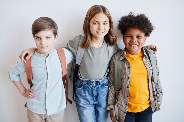 Photo portrait de taille de trois écoliers souriants portant des sacs à dos et regardant la caméra en se tenant debout contre le mur d'écriture