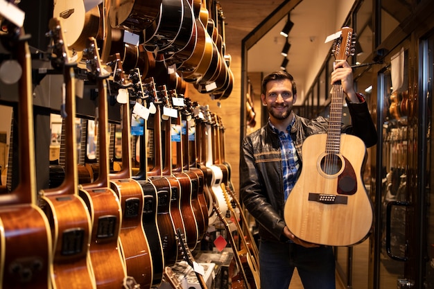 Portrait d'un talentueux musicien caucasien en veste de cuir debout dans un magasin de musique et tenant une guitare acoustique classique à vendre.