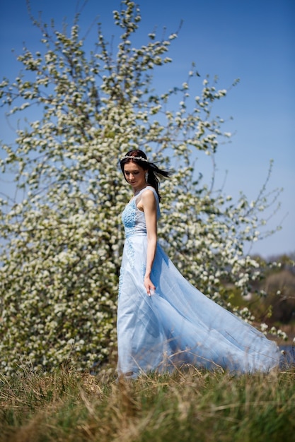 Portrait d'une tendre fille vêtue d'une longue robe bleue avec du tulle sous un cerisier en fleurs avec un sourire sur son visage par une chaude journée d'été ensoleillée