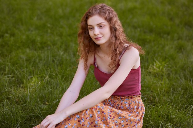 Portrait de tendre modèle paisible posant assis sur l'herbe, regardant droit devant, appréciant le repos après une longue marche, portant un haut rouge et une jupe colorée. Concept d'activités de personnes et de temps libre.