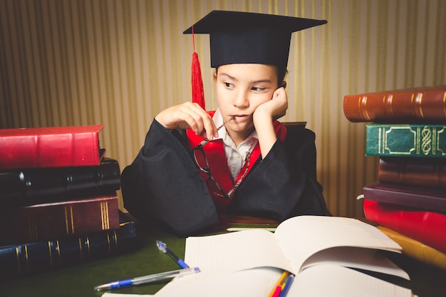 Portrait tonique d'une fille intelligente réfléchie en chapeau et robe de graduation au bureau