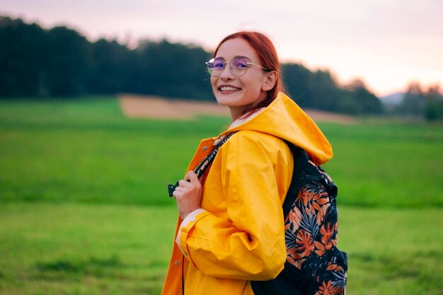 Portrait d'une touriste heureuse avec un sac à dos dans des verres portant un manteau jaune. Champ vert