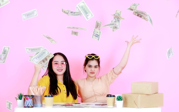 Portrait tourné en studio de deux femmes asiatiques riches et riches en démarrage, entrepreneur de petite entreprise, femme d'affaires partenaire assis ensemble jetant des billets de cent dollars pleuvant sur fond rose.