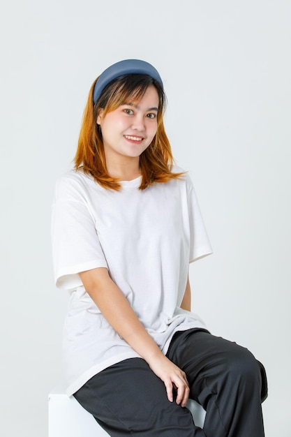 Portrait tourné en studio d'une jeune adolescente asiatique millénaire aux cheveux teints modèle féminin portant un bandeau et un beau maquillage en t-shirt décontracté et pantalon noir assis souriant regardant la caméra sur fond blanc.