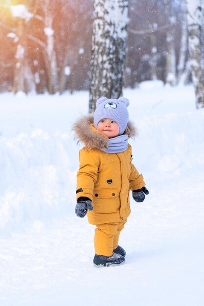 Photo portrait d'un tout-petit de 1520 mois dans des vêtements chauds jaunes regardant en arrière debout sur un chemin enneigé dans le parc lors d'une chute de neige