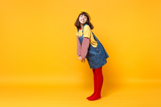 Portrait de toute la longueur d'une belle adolescente en béret français et robe d'été en denim debout isolé sur fond de mur jaune en studio. Concept de mode de vie des émotions sincères des gens. Maquette de l'espace de copie.