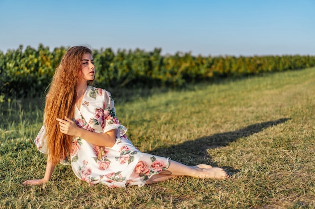 Portrait de toute la longueur de la belle femme aux longs cheveux bouclés sur le champ de cassis. femme dans une robe légère est assise sur l'herbe en journée ensoleillée