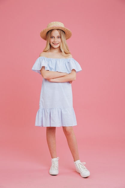 Portrait de toute la longueur de la belle fille blonde d'été 8-10 portant une robe bleue et un chapeau de paille posant à la caméra avec les bras croisés et sourire, isolé sur fond rose