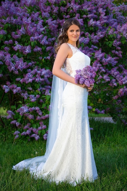 Portrait de toute la longueur d'une belle mariée avec un long voile debout près d'un lilas en fleurs dans un jardin d'été