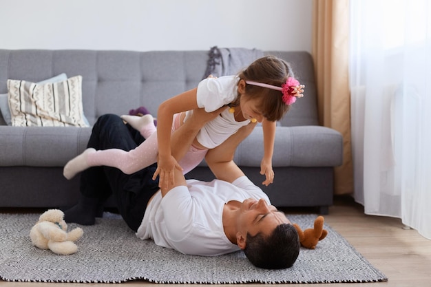 Portrait de toute la longueur du beau père brune jouant avec sa fille allongé sur le sol près du canapé, soulevant son enfant et souriant, portant un t-shirt et un pantalon blancs.