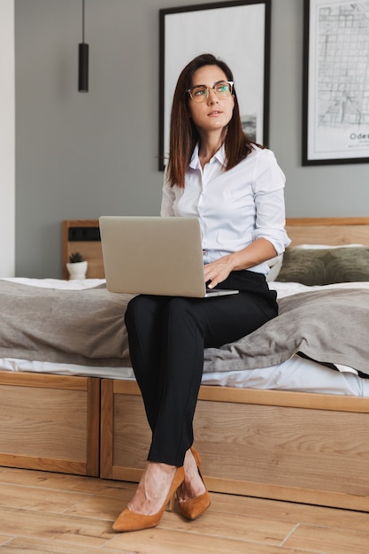 Portrait de toute la longueur d'une femme d'affaires adulte confiante en costume formel tapant sur un ordinateur portable assis sur le lit dans l'appartement