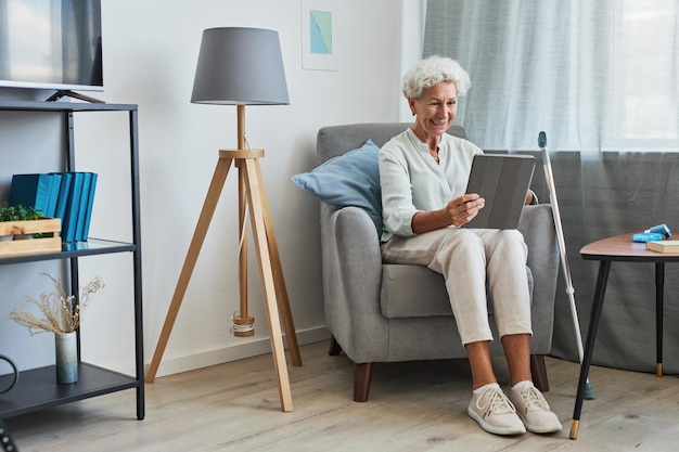 Portrait de toute la longueur d'une femme âgée souriante utilisant une tablette numérique à la maison tout en étant assise dans un confort...