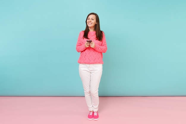 Portrait de toute la longueur d'une femme en pull rose tricoté, pantalon blanc envoyant des SMS sur un téléphone portable