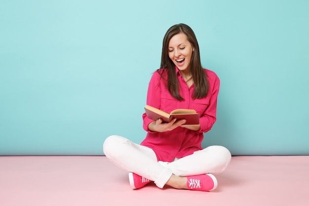 Portrait de toute la longueur d'une femme souriante en chemisier rose, pantalon blanc assis sur le sol lire le livre