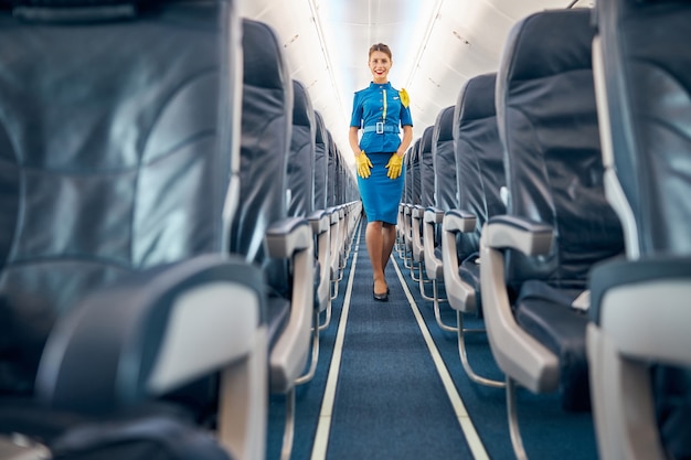 Portrait de toute la longueur d'une femme souriante debout près des confortables chaises de couleur sombre des passagers à bord d'un avion commercial