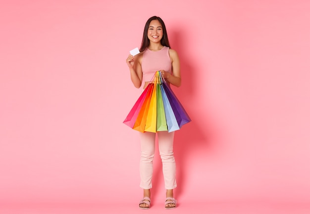 Portrait de toute la longueur d'une fille asiatique à la mode heureuse montrant sa carte de crédit et tenant des sacs à provisions, profitant de dépenser de l'argent sur de nouveaux vêtements, debout sur un mur rose.