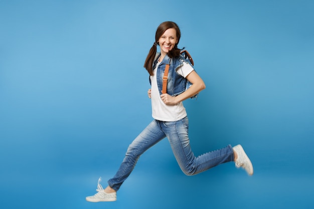 Portrait de toute la longueur d'une jeune étudiante drôle et souriante dans des vêtements en denim avec sac à dos sautant les jambes écartées isolées sur fond bleu. L'éducation au collège. Copiez l'espace pour la publicité.