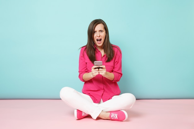 Portrait de toute la longueur d'une jeune femme amusante en chemise rose, un pantalon blanc assis sur le sol tient un téléphone portable