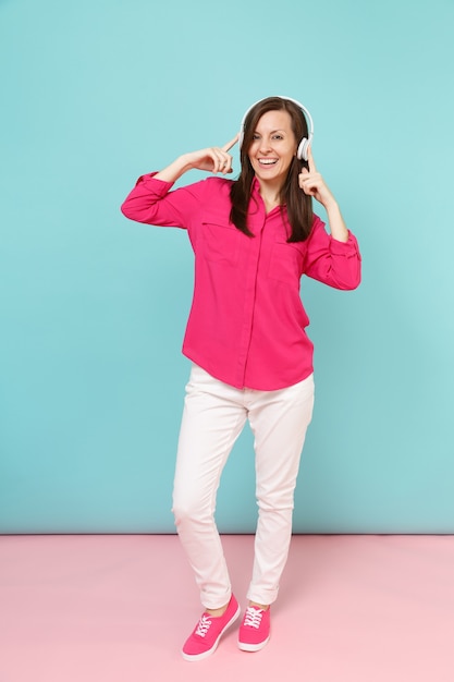 Portrait de toute la longueur d'une jeune femme amusante en chemisier rose, pantalon blanc, casque dansant isolé sur un mur pastel bleu rose vif.