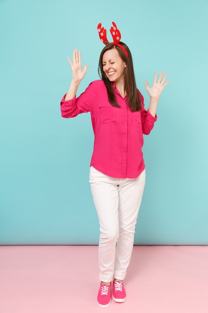 Portrait de toute la longueur d'une jeune femme amusante en chemisier rose, pantalon blanc, cornes de cerf posant isolées sur un mur pastel bleu rose vif.