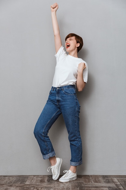 Portrait de toute la longueur d'une jeune femme brune criant et faisant des gestes en tant que gagnant isolé sur un mur gris