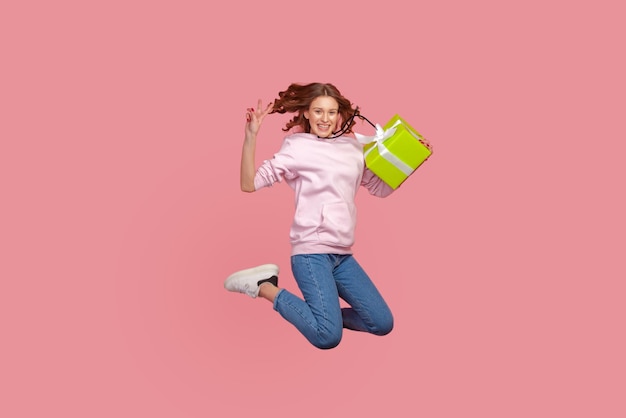 Portrait de toute la longueur d'une jeune femme excitée en sweat à capuche et jeans gesticulant signe de victoire et tenant une boîte-cadeau emballée en saut souriant sincèrement prise de vue en studio intérieur isolée sur fond rose