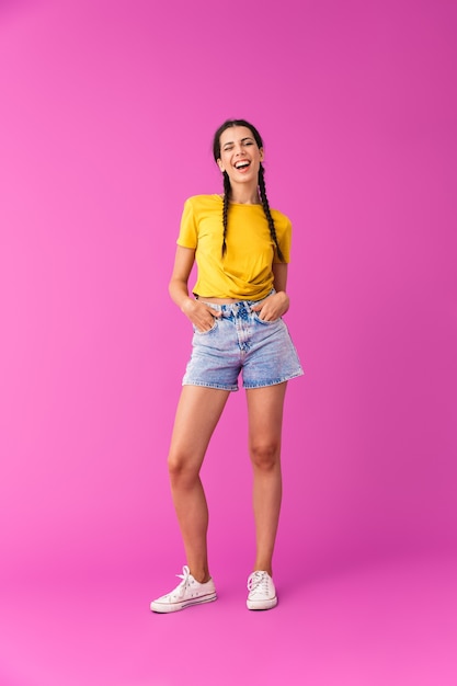 Portrait de toute la longueur d'une jeune femme joyeuse portant un short en jean clignant de l'œil avec ses mains dans les poches isolées sur un mur rose