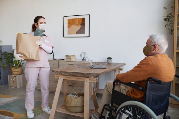 Portrait de toute la longueur d'une jeune femme tenant un sac d'épicerie tout en livrant de la nourriture à un homme âgé en fauteuil roulant, espace pour copie