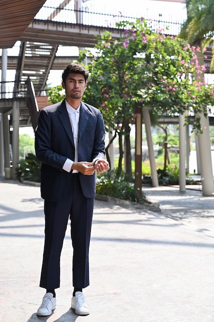 Portrait de toute la longueur d'un jeune homme d'affaires en costume noir formel à la mode en détournant les yeux tout en se tenant à l'extérieur près d'un immeuble de bureaux