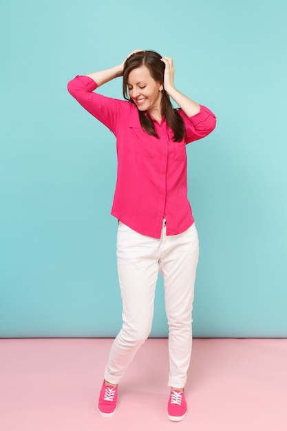 Portrait de toute la longueur d'une jeune jolie femme souriante en chemisier rose, pantalon blanc posant isolé sur un mur pastel bleu rose vif.