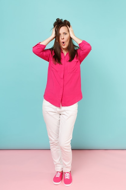 Portrait de toute la longueur d'une jeune jolie femme souriante en chemisier rose, pantalon blanc posant isolé sur un mur pastel bleu rose vif.