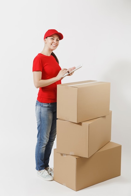 Portrait de toute la longueur de la livraison jeune femme au bonnet rouge, t-shirt isolé sur fond blanc. Courrier féminin debout près de boîtes en carton vides avec un ordinateur tablet pc. Réception du colis. Espace de copie