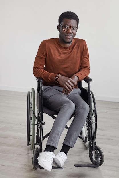 Portrait de toute la longueur minimale du jeune homme afro-américain utilisant un fauteuil roulant et tout en posant contre le mur blanc